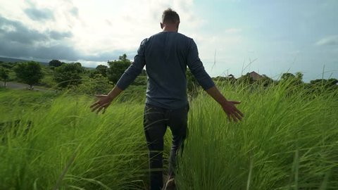 Camera follows man walks across tall lush green grass, touches grass with his hands. Shot from behind.