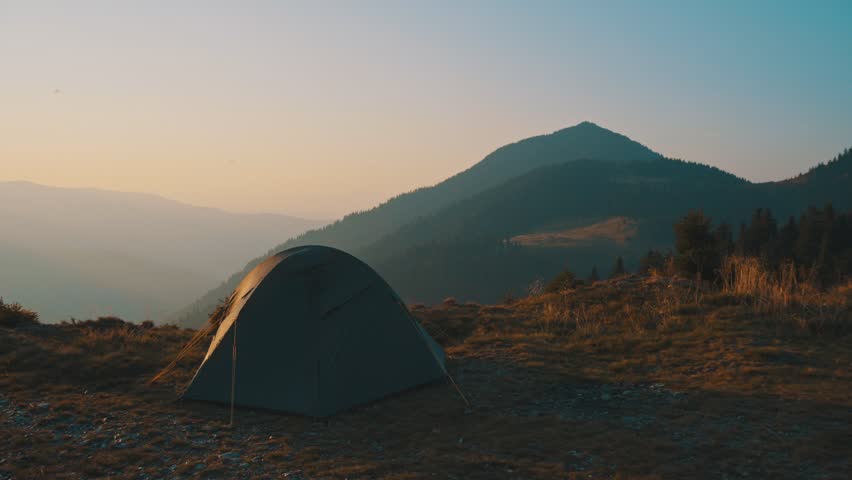 The male stands near the camping tent on the mountain on a sunset background