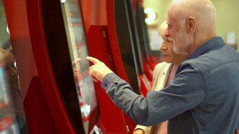 Senior couple choosing movie and buying ticket from vending machine at movie theater at mall via touch screen. Senior man making gestures by touching screen. Active modern life after retirement.