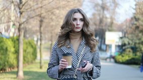smiling elegant woman in trench coat walking and drinking coffee from paper cup . smiling woman with curly hair look at camera smile walking slow motion face sunset beautiful lady outdoor closeup cute
