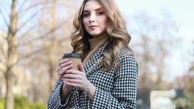 smiling elegant woman in trench coat walking and drinking coffee from paper cup . smiling woman with curly hair look at camera smile walking slow motion face sunset beautiful lady outdoor closeup cute