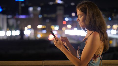 Young adult woman using smartphone at night, blurred city lights on background. Lady focused at phone, wear light summer dress at warm evening