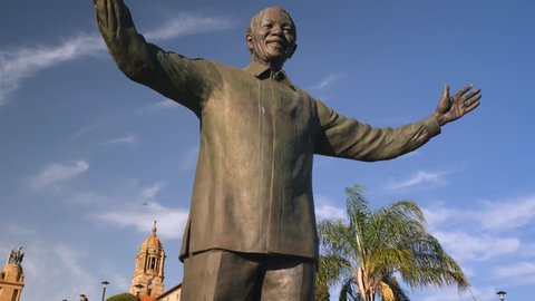 Pretoria, South Africa - circa 2019: Pull away from tall Nelson Mandela statue at government building, against blue sky. Tilt down, reveal garden, building behind, South African flag in wind.