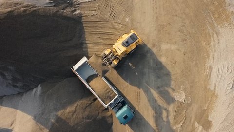 Top aerial view of bulldozer loading stones into empty dump truck in open air quarry.
