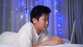 Happy preteen boy laughing while using laptop on bed with christmas light.