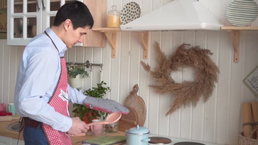 The young man cooks dinner, opens the lid of the pan, he is hot, he throws the lid and waves his hand.