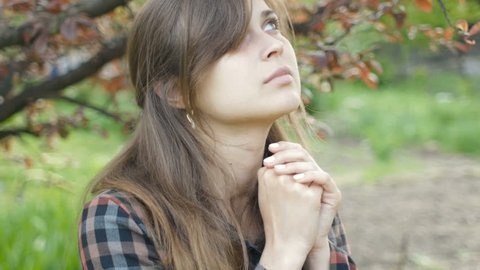 young woman turns to God with feelings in prayer, the girl folded her arms at her chest and with gratitude looks at the sky in the spring garden, the concept of religion and spirituality