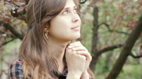 young woman turns to God with feelings in prayer, the girl folded her arms at her chest and with gratitude looks at the sky in the spring garden, the concept of religion and spirituality