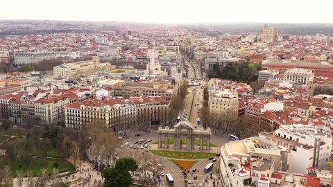 Busy Puerta de Alcalá in Madrid. Cars are driving on the street and pedestrians are entering and leaving the park El Retiro. The scenery is shown in an aerial shot on a semi cloudy day.