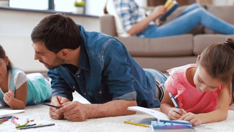 family, fatherhood and leisure concept - father spending time with his little daughters drawing in sketchbooks by crayons and lying on floor at home