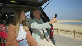 Couple photographing with smartphone in car. Beautiful happy young couple using smartphone while sitting in car at beach. Road trip concept
