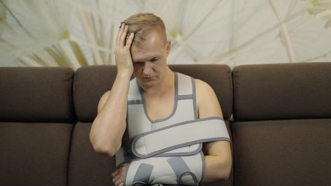 Man with shoulder injury. Painful, bored man with a broken arm wearing arm brace sitting on a sofa at home. Patient in a bandage for fixing of an elbow joint and a humeral belt. Slow motion