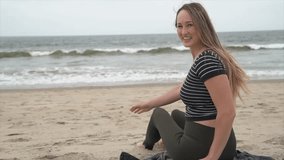 This slow motion video shows a young caucasian woman being silly, having fun, and laughing as she is throwing sand at camera while sitting on the beach.