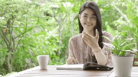 Young beautiful Asian woman video call her friends using high tech glass display in her home garden with green tree in background and drinking coffee. Future technology concept. For graphic overlay.