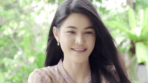 Super Slow motion of young beautiful Asian woman looking and smiling at camera in her home garden with green tree in background, close up shot. Millennial lifestyle concept.