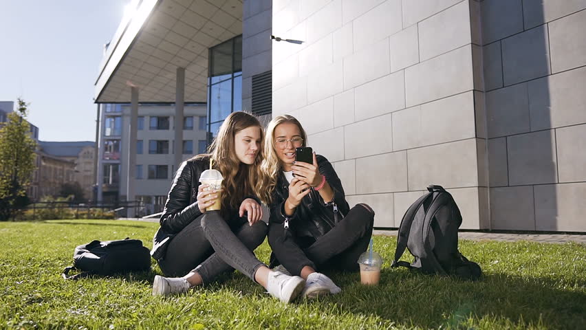 Attractive teen girlfriends with smartphone laughing at fun pictures through the internet sitting outdoors in the park in sunny weather. Outdoors. Slow motion