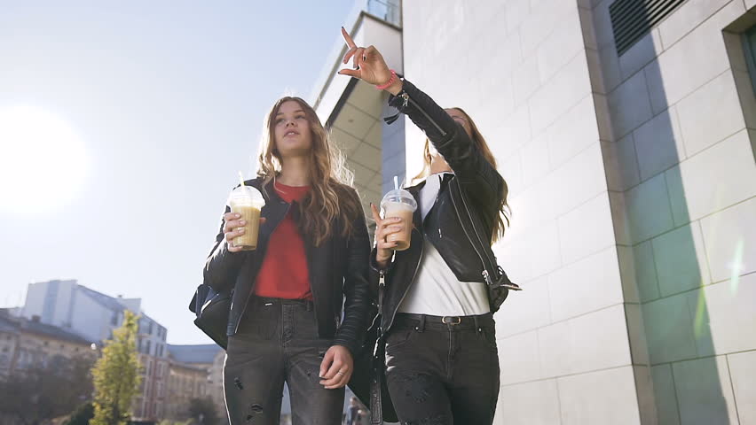 Two caucasian young female best friends drinking fresh juice while walking in the city near a modern building in sunny day. Outdoors, lifestyle. Slow motion