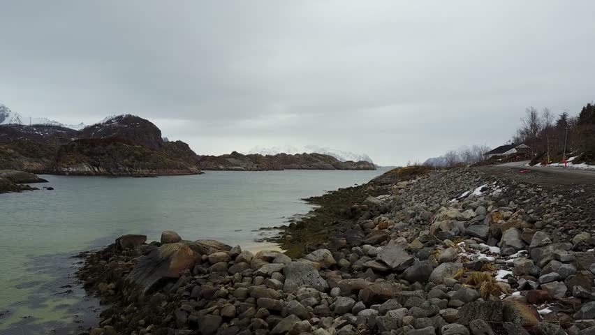 drone footage of shoreline rocks to cold river in winter.so quiet, moving forward like a cinematic movie into the middle of the water.snow mountain at distance with full grey clouds.norway in 4k 30fps