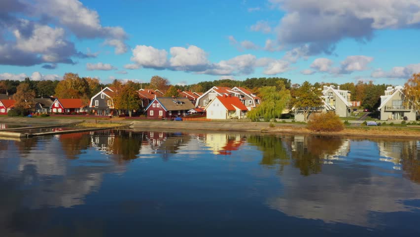 Forward rising drone shot of Pervalka, small village in Lithuania, surrounded by autumnal forest, near the coastline and the calm sea, reflecting white clouds on the surface, on a bright sunny day
