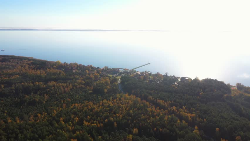 Drone shot of Pervalka, small village in Lithuania, surrounded by autumnal forest, near the coastline and the calm sea with reflecting surface, on a bright sunny day