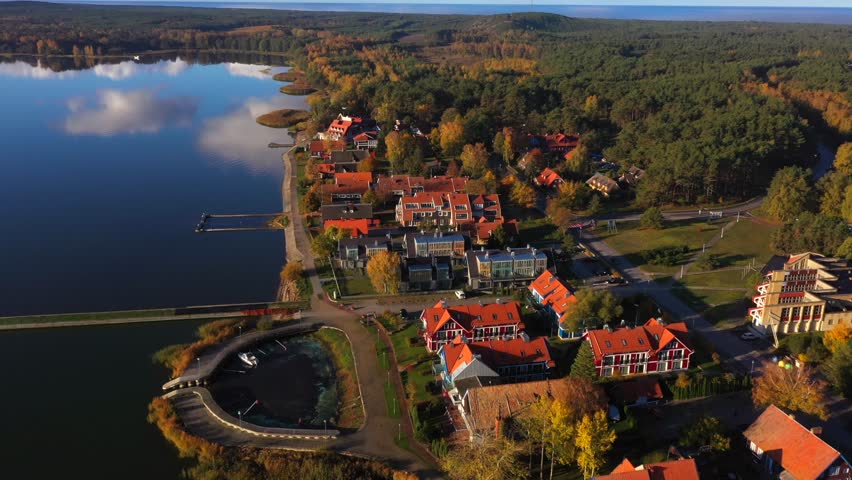 Drone shot of Pervalka, small village in Lithuania, surrounded by autumnal forest, near the coastline and the calm sea, reflecting white clouds on the surface, on a bright sunny day