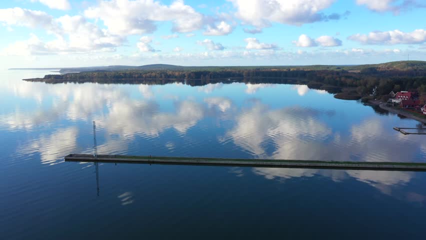 Panoramic drone shot of Pervalka, small village in Lithuania, surrounded by autumnal forest, near the coastline and the calm sea, reflecting white clouds on the surface, on a bright sunny day