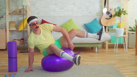 Funny young man with a mustache from the 80s trains and absurdly falls off a fitness ball in his house slow mo