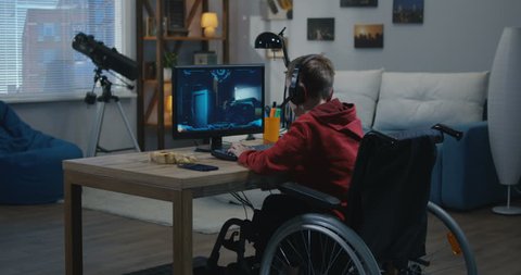 A medium wide shot of a disable young gamer playing video games online while sitting on his wheelchair