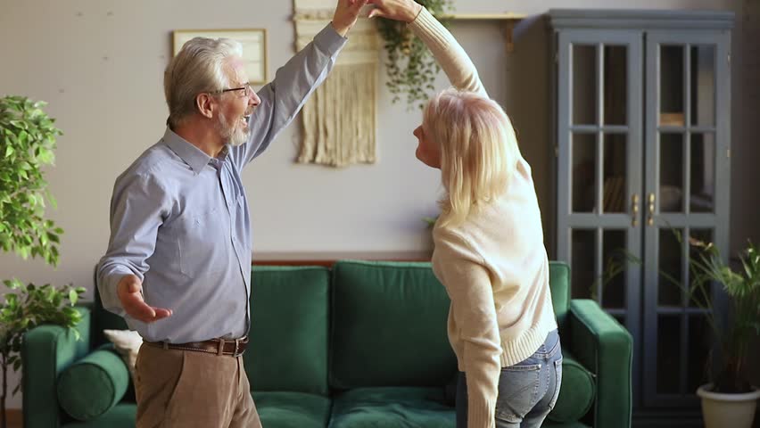 Happy old senior romantic couple dancing in modern living room, loving elder husband holding middle aged wife hand spinning around having fun at home, aged family grandparents relaxing together Royalty-Free Stock Footage #1028899871