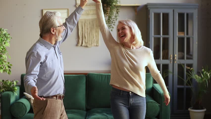 Happy old senior romantic couple dancing in modern living room, loving elder husband holding middle aged wife hand spinning around having fun at home, aged family grandparents relaxing together Royalty-Free Stock Footage #1028899871