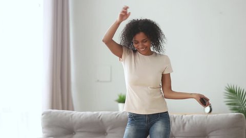Happy carefree african american millennial girl having fun at home listening to good music playing in smartphone app, funky energetic mixed race young woman holding phone dancing alone in living room