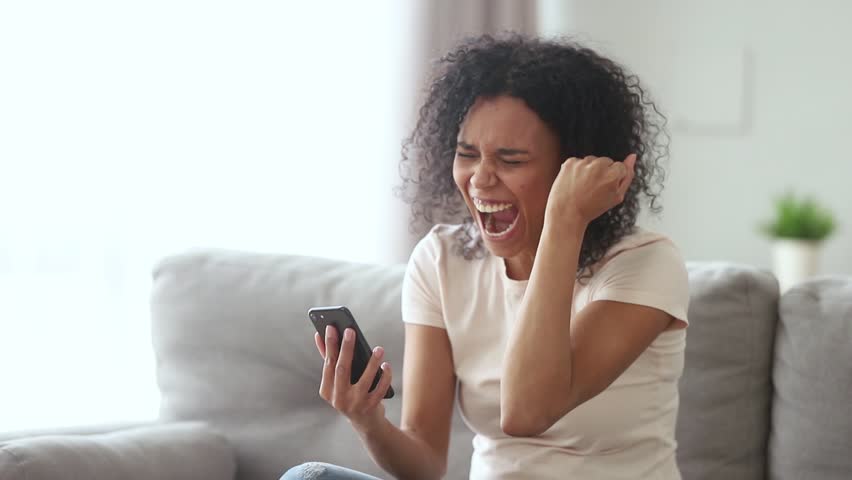 Happy young african american woman winner feel overjoyed celebrating bid win success victory looking at smartphone, excited black girl screaming yes winning online game app on cell phone at home