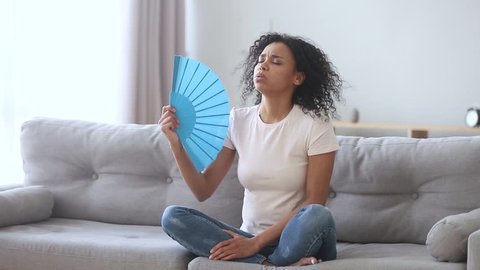 African american young woman feeling hot suffering from high temperature summer heat problem sitting on sofa at home, annoyed black girl holding waving fan sweating cooling in hot weather indoors