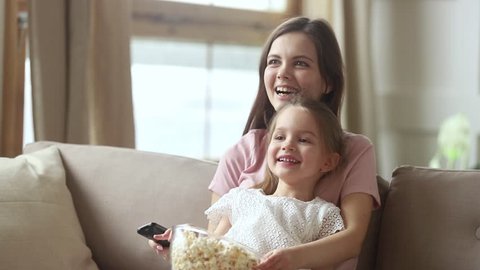 Cute little child daughter laughing sitting on young mom lap sofa eat snack popcorn enjoy funny television show, smiling mother holding remote control watching cartoons on tv with small kid at home