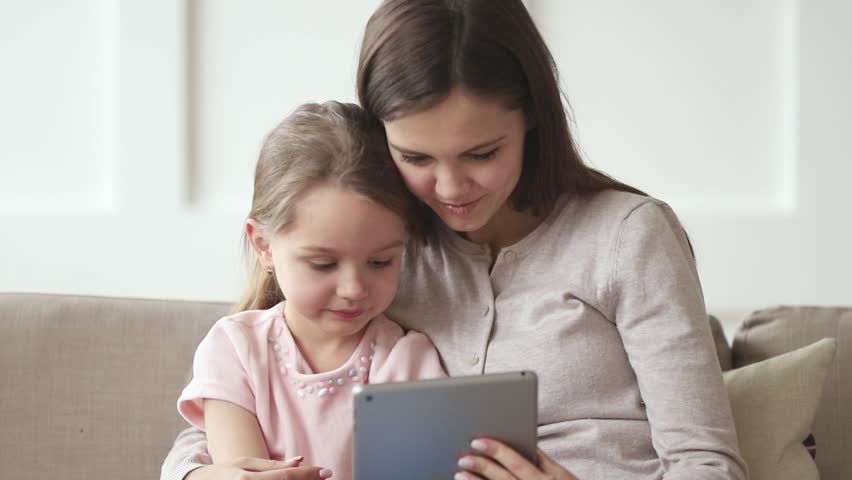 Happy family mom and kid daughter laughing having fun using digital tablet sitting on sofa, mother babysitter with child girl holding pc computer looking at screen watching funny cartoons online Royalty-Free Stock Footage #1028899961