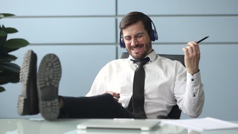 Happy funky businessman relaxing wearing wireless headphones listening to music pretending playing guitar at workplace, funny male worker enjoy favorite track having fun during work break in office