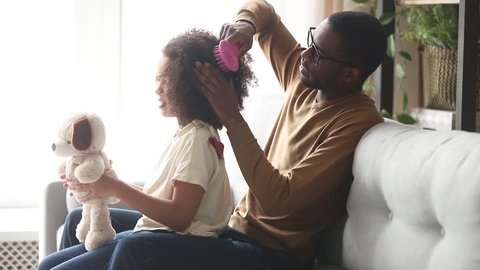 Loving black father doing brushing afro hair of cute little kid daughter talking bonding sitting on sofa at home, caring single african dad babysitter helping child girl with hairstyle in the morning