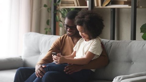Happy mixed race kid girl holding phone sit on african american dad lap on sofa, black father teaching little cute child daughter using apps looking at cellphone listen to music on smartphone at home