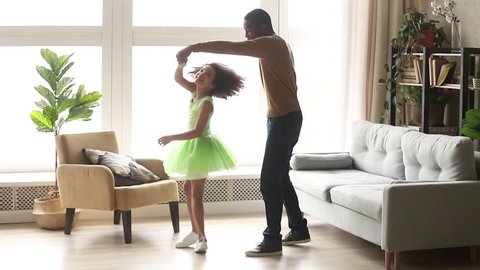 Happy african american dad dancing with child in dress playing spinning in living room, loving black dad stand on knee holding kiss hand of cute kid girl teaching daughter to dance having fun at home