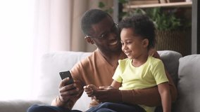 Happy african american dad and little son laughing looking at phone, black father teaching child boy using smartphone apps sitting on sofa, loving daddy holding cellphone talking to small kid at home