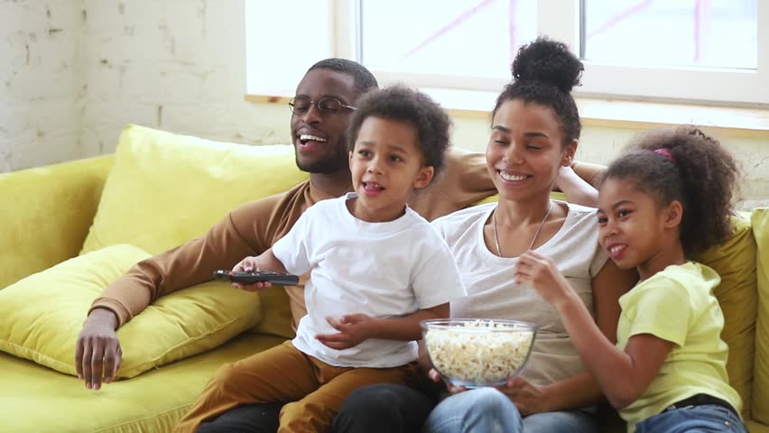Happy african american family mom dad and kids son daughter laugh hold popcorn remote control watch funny tv show sit on sofa together, black parents having fun with children view television at home Royalty-Free Stock Footage #1028900090