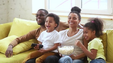 Happy african american family mom dad and kids son daughter laugh hold popcorn remote control watch funny tv show sit on sofa together, black parents having fun with children view television at home