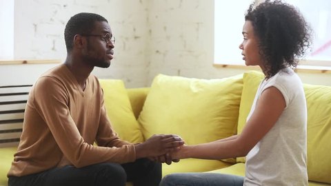 Young african american husband feeling guilt apologizing talking begging upset wife ask for forgiveness, black man holding hand of sad woman saying sorry please forgive get rejection sitting on sofa