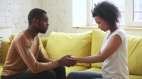 Loving african american husband holding hands of sad wife comforting giving empathy psychological support, caring black man consoling upset depressed woman having miscarriage sit on sofa at home