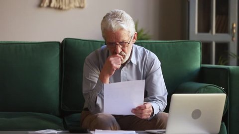 Serious old retired man holding paper calculating domestic bills at home using calculator laptop, senior aged grandfather counting household payments to pay online on computer sitting on sofa alone