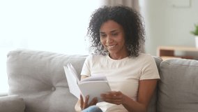 Smiling young african american woman sitting on sofa at home holding enjoying reading open book, happy mixed race girl relaxing spending time with modern literature on couch at home alone on weekend