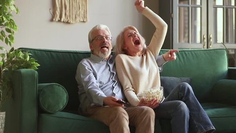 Overjoyed old mature couple football fans watching sport tv game support cheer winning soccer team sit on sofa together, happy excited senior family celebrate victory goal score view television match