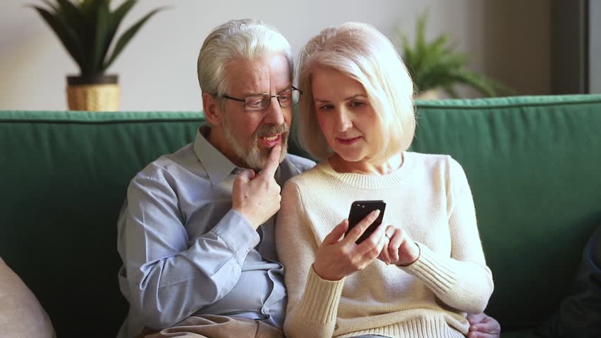 Happy old mature retired couple holding smartphone looking at cellphone screen laugh watching funny photos sit on sofa, cheerful senior family having fun enjoy read online news in smartphone at home Royalty-Free Stock Footage #1028900204