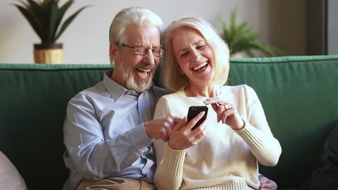Happy old mature retired couple holding smartphone looking at cellphone screen laugh watching funny photos sit on sofa, cheerful senior family having fun enjoy read online news in smartphone at home