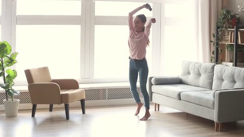 Happy carefree young woman dancing alone in modern living room with big window listen to music on smartphone, funky millennial girl holding phone enjoy new hit song playing in app having fun at home